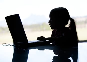 A young girl sits at a laptop and is blacked out and positioned side on