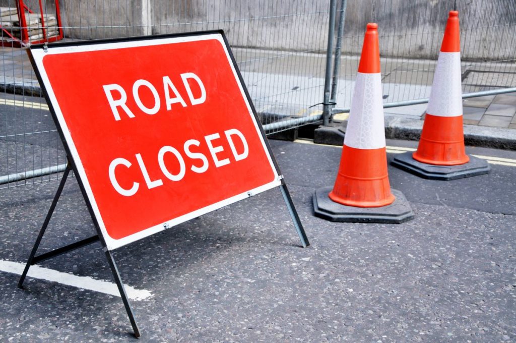 A road closed sign accompanied by two orange and yellow traffic cones