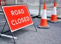 A road closed sign accompanied by two orange and yellow traffic cones