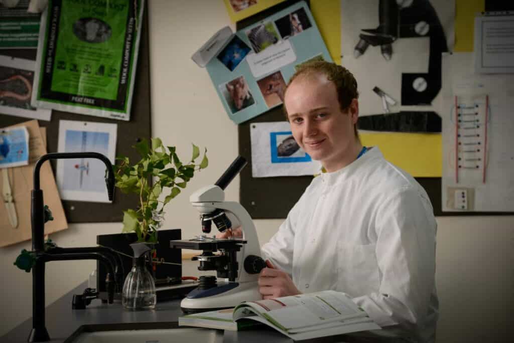 Toby Wilson is pictured in a laboratory with a microscope and textbook. He is smiling and has ginger, balding hair