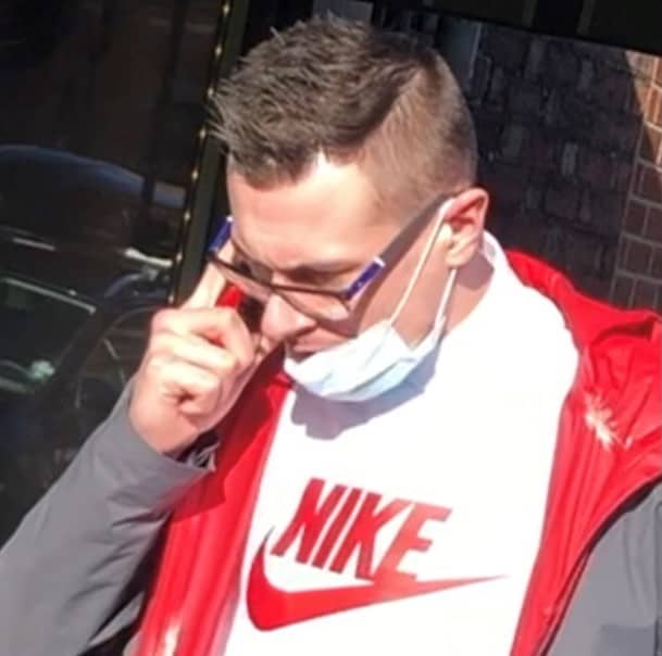 Aidan Smillie is holding his phone to his ear and is wearing glasses and brown, spiked hair. He is wearing a white Nike t-shirt with a red Nike logo with the Nike tick. He is wearing a face mask that was not covering his face or mouth. He had a grey hoodie with a red lining over his t-shirt