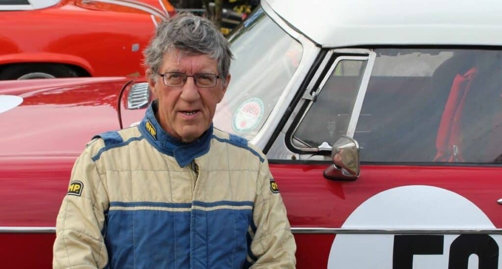 Graeme Forrester pictured with a red car in the background. He is wearing glasses, a white and blue race suit and has grey hair. 