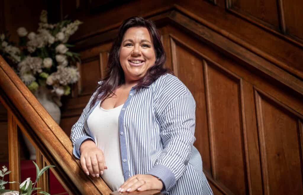 Rachael Bell Wealth Management Practice Principal stands with her arms on a stair bannister with a smile on her face. She is brunette and has a white and blue striped shirt which is open with a white t-shirt underneath