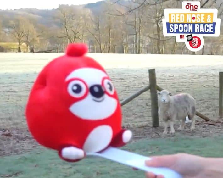 A screenshot from a promotional video for The One Show Red Nose & Spoon Race for Comic Relief. It shows a red nost balanced on a spoon with the Lake District and a sheep in the background