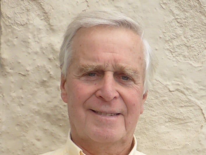 Ron Palmer smiling against a white wall. He has grey hair and is wearing a yellow shirt 