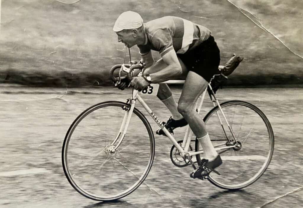Albert White on his way to team gold in the 1961 National Time Trial. The black and white image shows him cycling with a white cap and a striped polo shirt