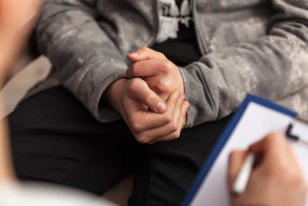 A young person holds his hands together in a nervous position while speaking to a counsellor. The youngster is wearing a grey hoodie and black trousers