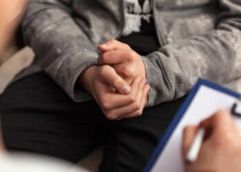 A young person holds his hands together in a nervous position while speaking to a counsellor. The youngster is wearing a grey hoodie and black trousers