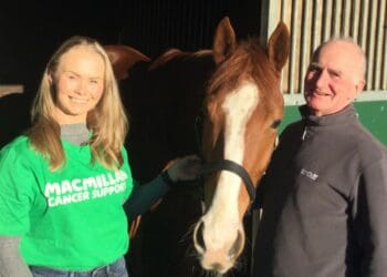 Amateur rider Morgan Prescott is pictured wearing a green Macmillan t-shirt and grey jumper underneath. She has blonde hair and is smiling. Behind her in the stable is a brown horse with a a white stripe down the front of its head. On the right is racehorse owner James Callow. He is wearing a grey fleece. He has grey balding hair