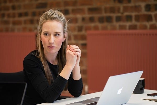 Helen Statham, Intro co-founder and director pictured with her hands together while leaning on a table. She is sitting with Mac laptop and is wearing a black long-sleeve blouse. She has blonde hair