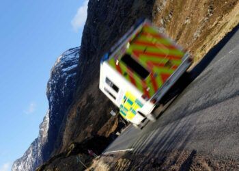 An ambulance travels on a rural road with the fells in the background
