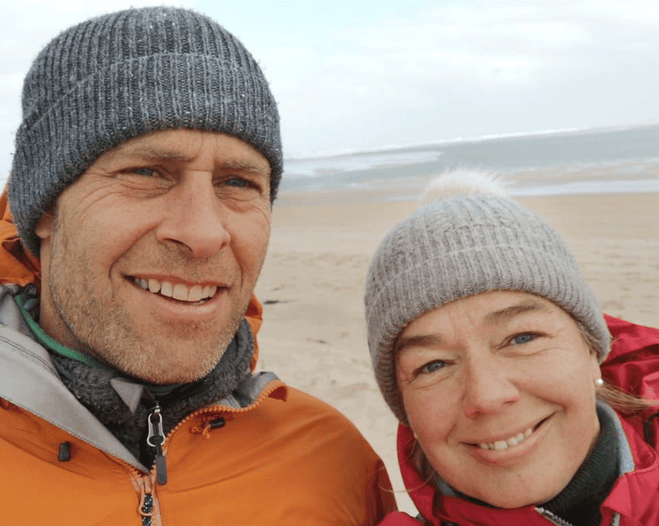 Tom Horne is pictured with a grey woolly hat and orange jacket. Lesley (on the right) is wearing a white woolly hat and a red jacket. Both are smiling and are on the beach