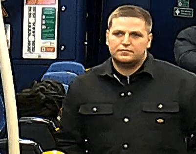 The man pictured has short mousey brown hair. He is wearing a black coat with buttons in the aisle of a train
