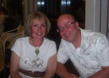Moira is pictured on the left with blonde, shoulder-length hair and with a smile on her face. She s sitting down and leaning into Gary with a white t-shirt and brown belt. Gary is bald with glasses and is also leaning in towards Moira. He is wearing a white shirt and is also smiling