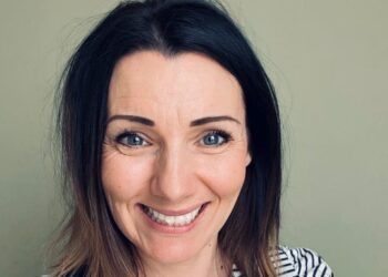 Rebecca has shoulder-length brown hair and is smiling. She is wearing a black and white striped top and black dungarees