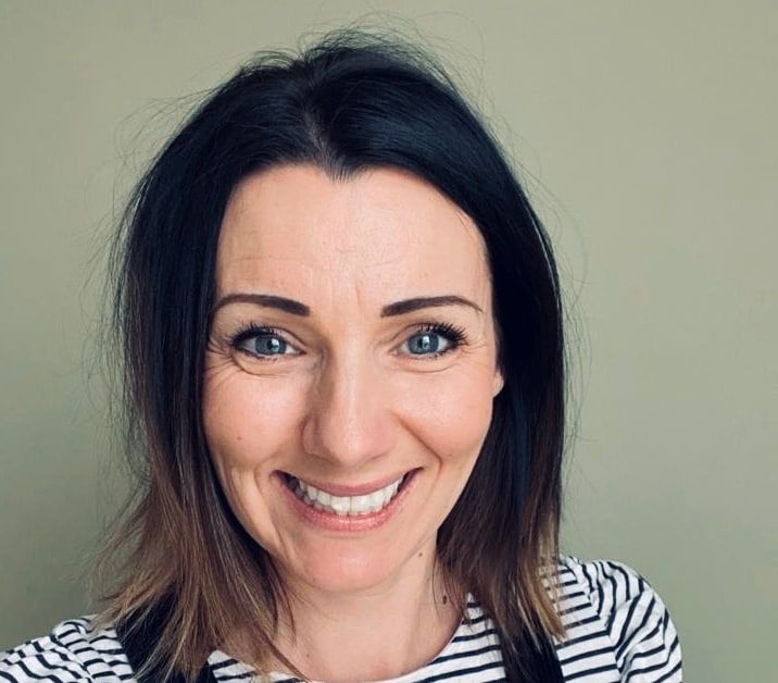 Rebecca has shoulder-length brown hair and is smiling. She is wearing a black and white striped top and black dungarees