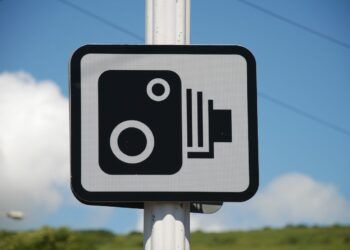 A sign indicating CCTV camera surveillance, black and white graphic on a white background with a black border, mounted on a post outdoors with a blue sky and green grass in the background.