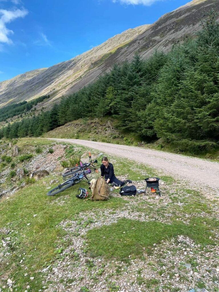 Alex cooking sausages for Apollo in Ennerdale with a backdrop of mountains and pine trees