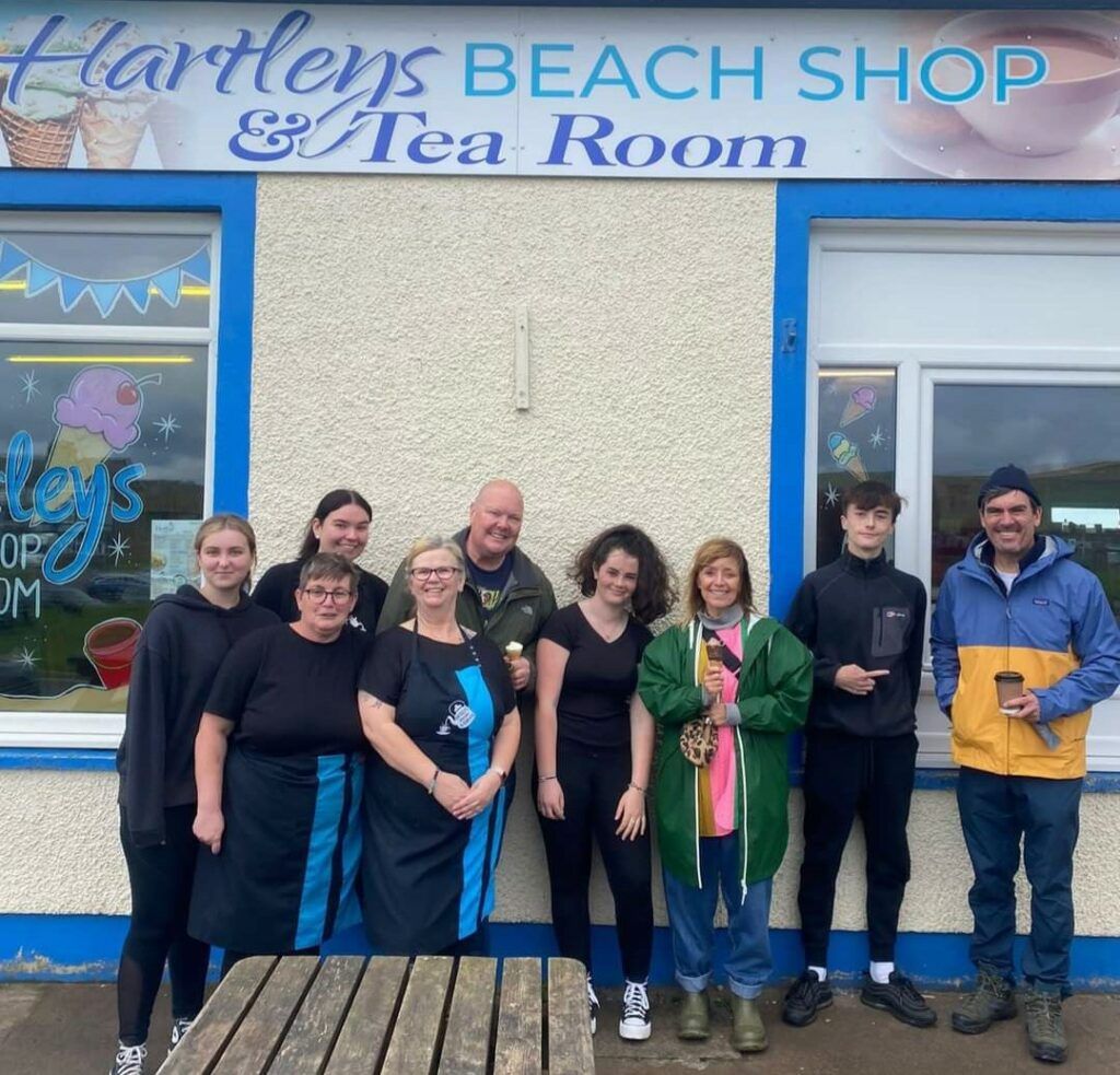 The St Bees Hartley's team with Emmerdale's Paddy, Cain and Rhona stood outside the front of the tea room smiling with the tea room's sign in the background 