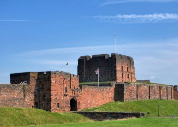 A historic castle with red brick walls and towers set against a blue sky with green grass in the foreground, showcasing medieval architecture.
