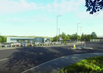 A motorway service station with cars and lorries parked outside, surrounded by trees and greenery under a blue sky.
